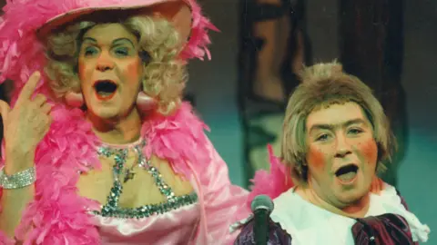 King's Theatre Walter Carr and Gregor Fisher performing onstage - Carr is playing a panto dame with a pink dress and large pink hat on. Fisher is also in a purple dress. 
