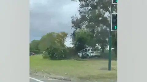Louise Lefevre The white car on the roundabout. It has hit a tree, which is damaged. The car is also badly damaged. It is covered in black scorch marks and its windows are smashed.