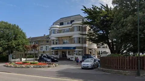 A four-storey art deco style hotel stands behind a small circular flower bed, with several cars parked in front of the building. Large green trees stand on both sides of the building