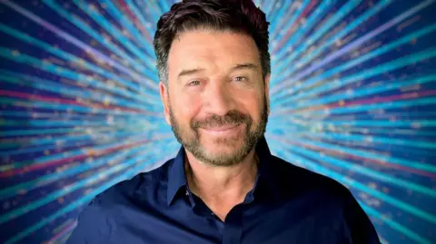 BBC Nick Knowles, a antheral   with acheronian  hairsbreadth  and stubble, wears a acheronian  bluish  garment  and stands against a bluish  starburst background, smiling 