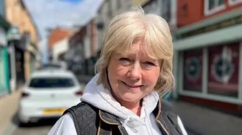 Sarah Jane Watson is the Managing Director of Visit Cleethorpes which is an online platform which promotes tourism to the area. She has short blonde hair and is smiling into the camera. Sarah Jane is wearing a grey top with a hood along with a dark coloured padded body warmer. She is stood on Sea View Street in Cleethorpes which has a range of independent shops and a cafe culture.