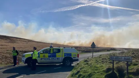 A police 4x4 vehicle parked across a road with smoke billowing in the background across open moorland