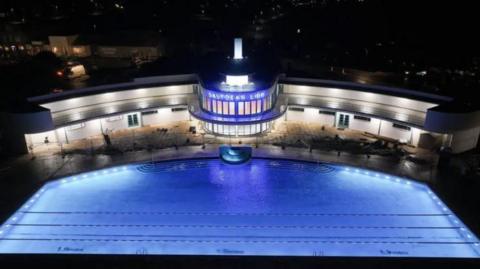 Saltdean Lido's illuminated blue outdoor swimming pool pictured from the air at night with the large curved white lido building and neon sign lit up in the background 