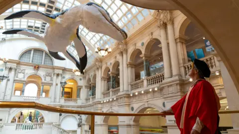 University of Bristol A giant model of a pliosaurus is suspended over a large grand building. A man wearing a red graduation robe is standing on a glass balcony looking up at the pliosaurus.