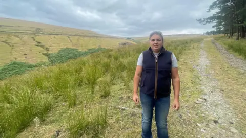 A woman in a blue bodywarmer, blue jeans and a grey t-shirt stood on a large countryside moor.