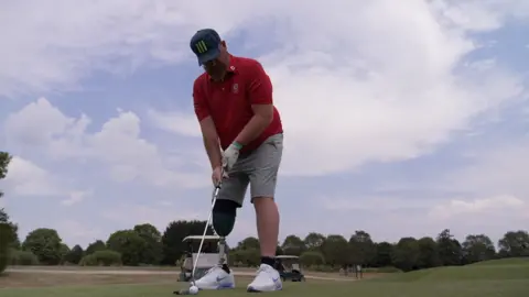 BBC A man with a prosthetic leg stood with holding a golf club about to hit a golf ball.