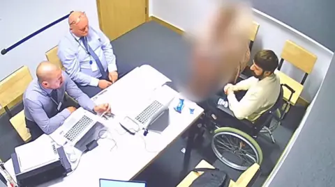 Hertfordshire Police Police interview image of Clifford, who has dark hair and a beard, a white jumper and in a wheelchair, speaking across a table to two male officers who are both wearing purple shirts and ties and sat with laptops in front of them.