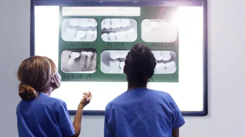 Getty Images Two dental students in blue scrubs with their backs to the camera, looking at a screen with several x-rays of sets of teeth.