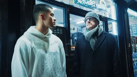 Netflix Louis Theroux, dressed in a heavy coat and woolen scarf and hat, speaks to influencer Sneako outside a Bodega (corner shop) in New York City. The bright lights from inside the shop illuminate the street where the men stand. Sneako, who wears a white hoodie with a faint gray design on the front, is looking sideways at Theroux during an apparent pause in their conversation.