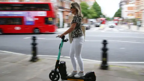 Getty Images A blonde woman wearing white jeans, white sneakers, a leopard print top and sunglasses riding a green and black electric scooter 