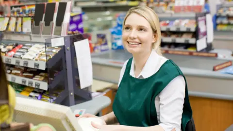 A blonde haired shop worker with a white top and green, sleeveless vest (uniform), she is sat beside a till, smiling and scanning products