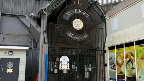 The entrance to Barrow Market Hall which has a black fence in front of it. The roof is pointed and there is a large circular sign towards the top with a coat of arms and old lettering which reads 'Market Hall'. There is a sign stuck on the gate which has black and yellow tape surrounding it. There is a Herons shop to the right which multiple pictures of food on the side of the wall.