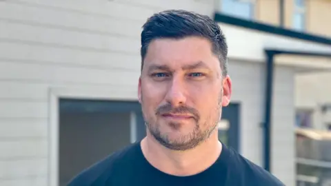 Stephen Huntley/BBC Gavin Crane is standing in front of a house which its exterior is made of grey wood. He has short brown hair and moustache and is wearing a black T-shirt.