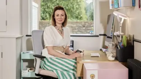 Handout Gee is sitting at a study desk with a flat-screen computer, a tray of pens and pencils and a large glass egg-timer. She is smiling to camera and wearing a white blouse and a green and white-striped skirt. There is a large window behind her with a brick wall and a tall hedge.
