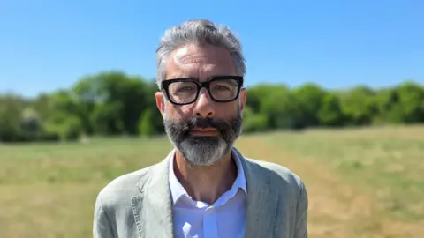 Paul Moseley/BBC Asher Minns is wearing a light linen suit jacket over a white shirt. He's wearing thick rimmed glasses.