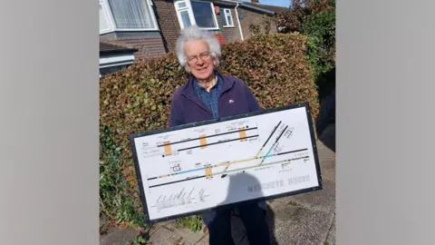 Network Rail A member of the Northumberland Line Heritage Community Group holding a diagram from Marcheys House. The diagram shows lines representing the track. They changre from black to green, blue and yellow. The man holding he diagram is wearing a fleece and has grey hair.