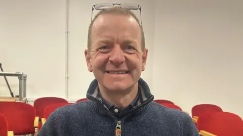 Robbie Jones smiling. He has light close cropped hair and his glasses are pushed up over his forehead. He is wearing a blue-grey quarter zip jumper.