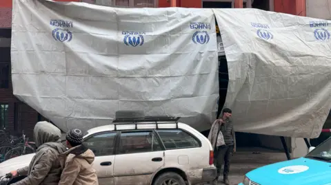 White sheets hang from from a building painted orange. A white car sits in front of the sheets, and people are in the street.