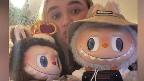 Ashley Bushey a woman with brown hair is looking directly at the camera and posing with two Labubu dolls. One is wearing a hat and the other is brown with a crown on top of its head. 