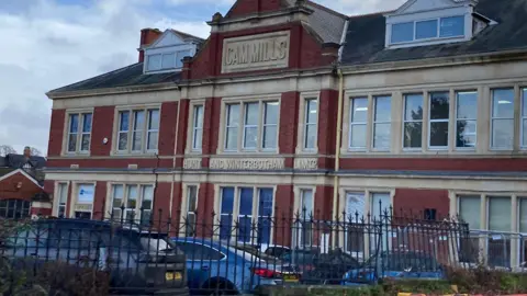 The outside of the Cam Mills building where WSP Textiles works operates from. There is a black fence in the foreground with parked cars in front of it.The building is red brick with sand stone around its windows. 