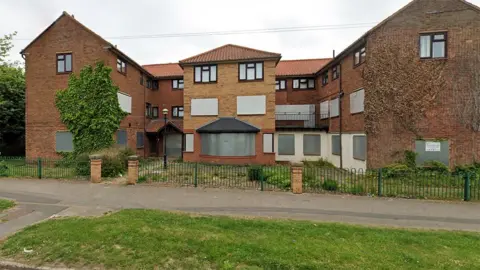 Google Exterior of Charles Brady Court it is a modern three-storey brick building with a pitched roof and a forecourt of grass behind green fences. All the windows and doors have been boarded up with metal panels