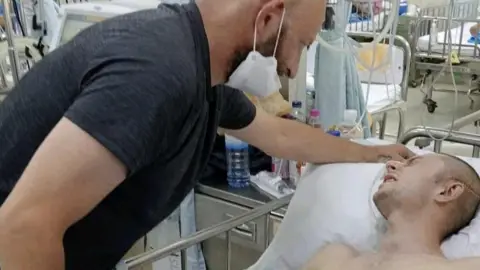 Family photo A man with a shaven head lies in a hospital bed. Leaning over him is a bald man wearing a black t shirt and a white face mask. Behind them is a table with various drinks and medical supplies on it and behind that are more rows of hospital beds.