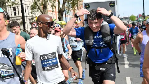 Jordan Adams wearing a blue t-shirt and black running shorts runs with a white fridge on his back surrounded by other runners. Mo Farah, wearing a white t-shirt and sunglasses, runs beside him.