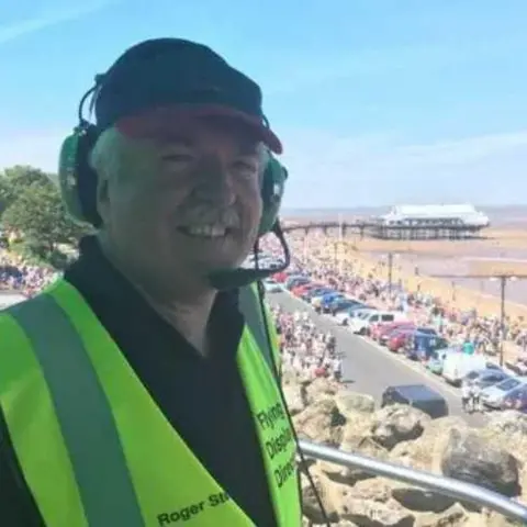 North East Lincolnshire Council A man with white hair and a matching moustache smiles as he stands on a gantry above a beach packed with people. He is wearing a black cap, a headset and a high-vis yellow jacket over a black polo shirt. 