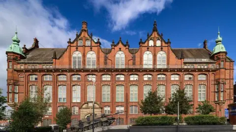 Museos del Noreste. El Museo del Descubrimiento es un edificio victoriano de ladrillo rojo con altos ventanales en su sección central. Dos torres verdes en forma de cúpula se alzan en cada extremo. Hay un tanque frente a él, césped bien cuidado y árboles jóvenes. El cielo es azul con algunas nubes dispersas.