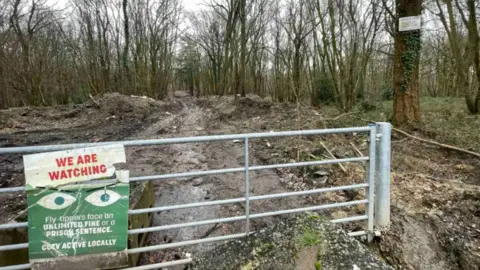 BBC A silver gate on a path into Hoads Wood with a sign on saying: 
WE ARE WATCHING
Fly tippers face an unlimited fine or a prison sentence.
CCTV ACTIVE LOCALLY