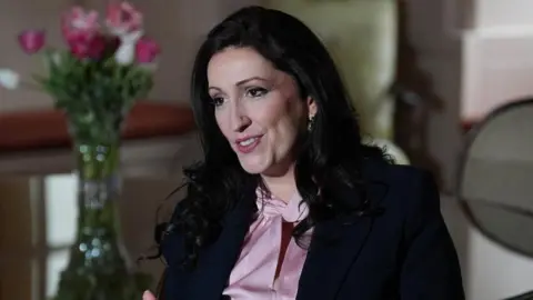 Emma Little-Pengelly, sitting on a chair, wearing a pink shirt and a navy blazer. She has long black hair. There is a table with a vase of flowers on top of it behind her. 