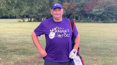 Tom Morris, Captain of the New Forest Golf Club, is stood wearing a purple Abby's Heroes t-shirt and cap, smiling at the camera while holding his golf clubs on one of his shoulders. He is stood on a golf course and is wearing a white golf glove on his left hand.