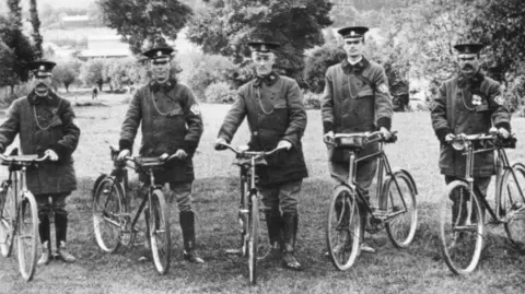 The AA A black and white photo of five AA patrolmen from 1914 wearing uniform of long jackets and knee-high boots and caps, standing next to their bicycles in a field