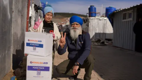Khalsa Aid A child, wearing a hat and a fleece, and a man wearing a blue turban and a navy blue fleece are posing by some boxes.
