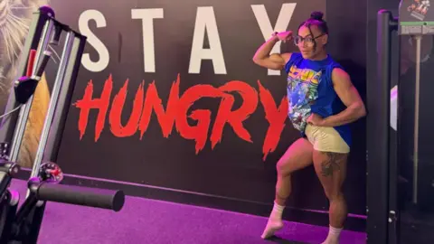 Kingdom Gym A woman in blue t-shirt and yellow short cycling shorts poses with her bicep curled, showing large muscles, in front of a wall banner which says in white and red Stay Hungry. Gym equipment can just be seen in the foreground. The woman has dark hair tied back and is wearing clear-framed glasses.