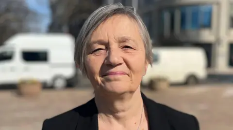 Mary is standing outdoors in a paved urban area, facing the camera. She is wearing a dark jacket over a patterned top. In the background are light-colored buildings, trees without leaves, and parked vans under bright daylight.