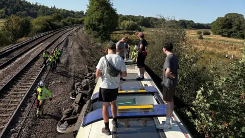 Alvin Dybvik Payne Four people stood on top of an overturned train carriage next to a train track.