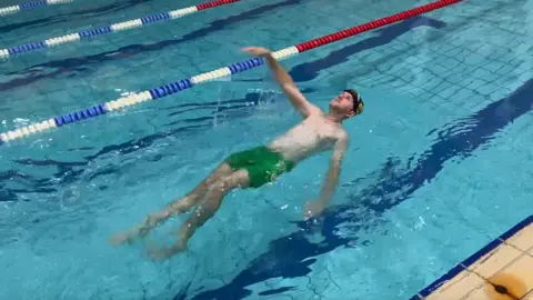 Michael Coombes, wearing green swimming shorts and doing backstroke in a pool.