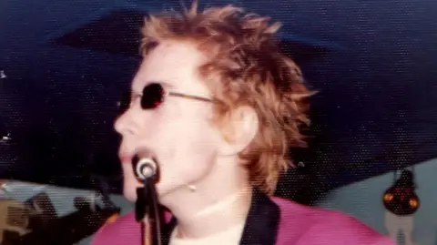Cindy Stern / Restoration by Tim Naylor A close-up image shows a performer with short, spiked hair singing into a microphone on stage. The performer is wearing dark sunglasses and a bright jacket, giving a bold, distinctive style.