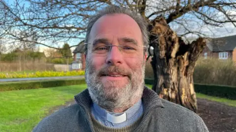 Rev Cawthorne looking into the camera, he's wearing a grey jumper and you can see he's wearing a vicar's clerical collar underneath. Behind him you can see some of the affected homes on Abbott Drive, and to his right is an oak tree.