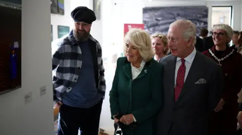 Reuters King Charles III and Britain's Queen Camilla meet photographer Cathal McNaughton during a visit to the Roe Valley Arts and Cultural Centre, in Limavady. They are all looking at a photo on the wall. The king has white hair and is wearing a dark grey suit, white shirt and red polka dot tie. The queen has blonde hair and is wearing a green skirt suit. Cathal McNaughton is on the left, he has a dark beard, wearing a black flat cap, blue and white check shacket, light blue t-shirt and blue jeans.