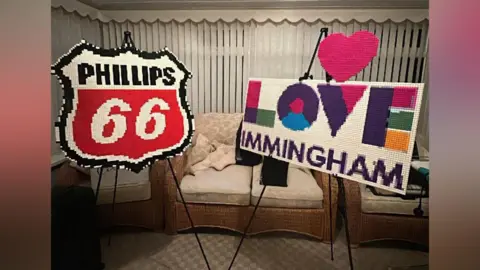 Brad Charlton-Wells Two large Lego builds of signs that are on black stands. On the left is a shield shaped sign that is white with a black outline. In the middle in black writing it reads "Phillips". Beneath is a red section with the number 66 in white writing. On the right is a large rectangular sign that reads "LOVE IMMINGHAM". The word love is written in purple, pink, green and yellow bricks. The word Immingham is in purple bricks. Above the sign is a pink lego heart