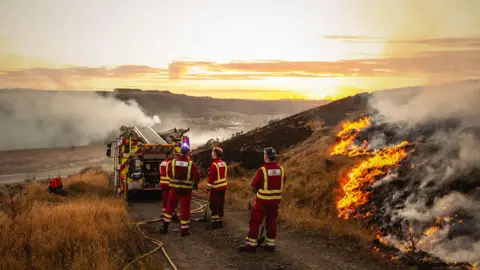Firefighters are stood next to very dry grass, which some if it has have been burnt or is on fire. A fire truck is pictured on a path, and the countryside in the distance has smoke coming from the ground.