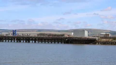 Billy McCrorie A pier with a railway station next to it and water in front and rolling hills in the background