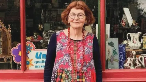 Contributed Marcia Riddington, with short red hair and black round glasses stands in front of shop front. She is wearing a dress with long black sleeves and the body is red with a floral pattern. 