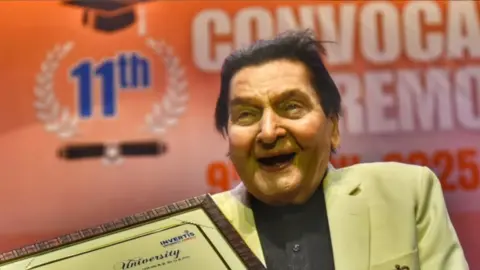 Asrani/Instagram Asrani holding a certificate and wearing a beige jacket with a black shirt is seen laughing