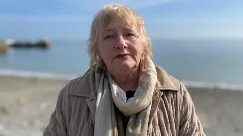 Georgina O'Sullivan is an older lady with short to medium light blonde hair. She is wearing a cream scarf and a beige jacket, standing in front of a stone beach on a sunny day.