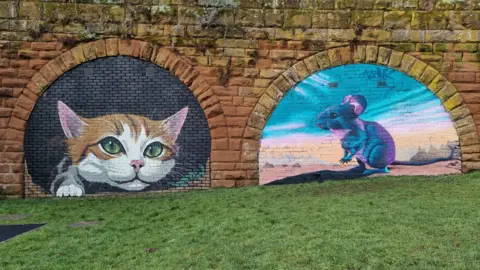 Tim Robottom Two filled in brown brick archways that have been painted on. The mural on the left is a ginger and white cat that appears to be climbing out of the bricks. It has green eyes and there are some leaves painted next to it. On the right is a painting of a grey mouse, with light shining on it. The mouse is standing in a desert-like landscape with hills and rocks in the background.