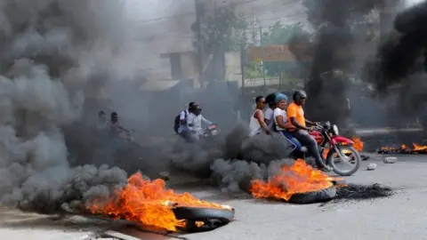 Reuters Motorcyclists with passengers drive past burning tyres in Port-au-Prince, Haiti. Photo: 13 July 2022