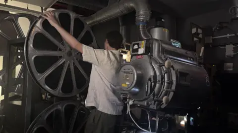 Patrick stands with his back to the camera, operating a large projector. He has one hand outstretched to the reel, and the other checking the mechanical operation of the machine. The room is dim and full of equipment, with lots of wires and large silver pipes. 